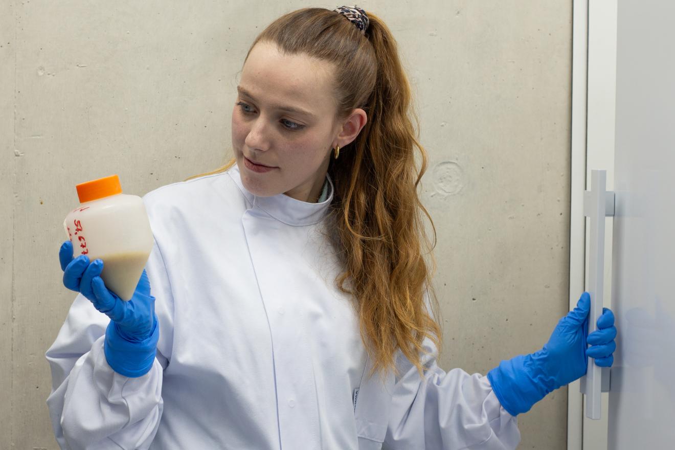 Eine Frau im Laborkittel betrachtet ein Gefäß mit Biomasse.