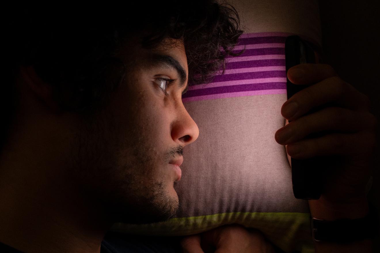 Ein junger Mann liegt mit dem Kopf auf einem Kissen mit violetten Streifen und blickt nachdenklich zur Seite. Die Szene ist weich beleuchtet und zeigt nur sein Gesicht im Profil.