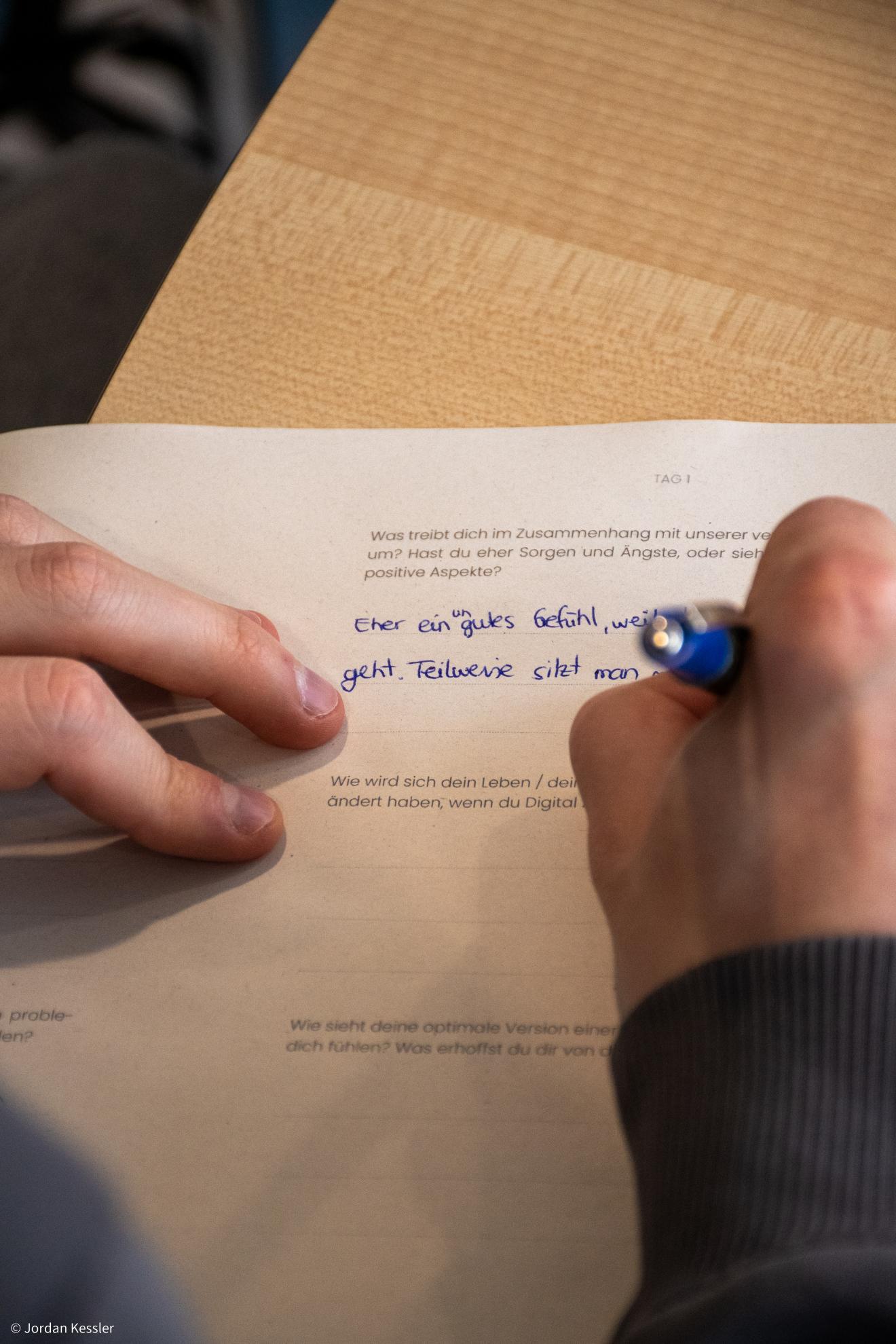 Eine Person schreibt mit einem blauen Stift auf ein Arbeitsblatt eines Workshops. Zu sehen ist eine Frage zum Thema digitale Medien und persönliche Gefühle dazu.
