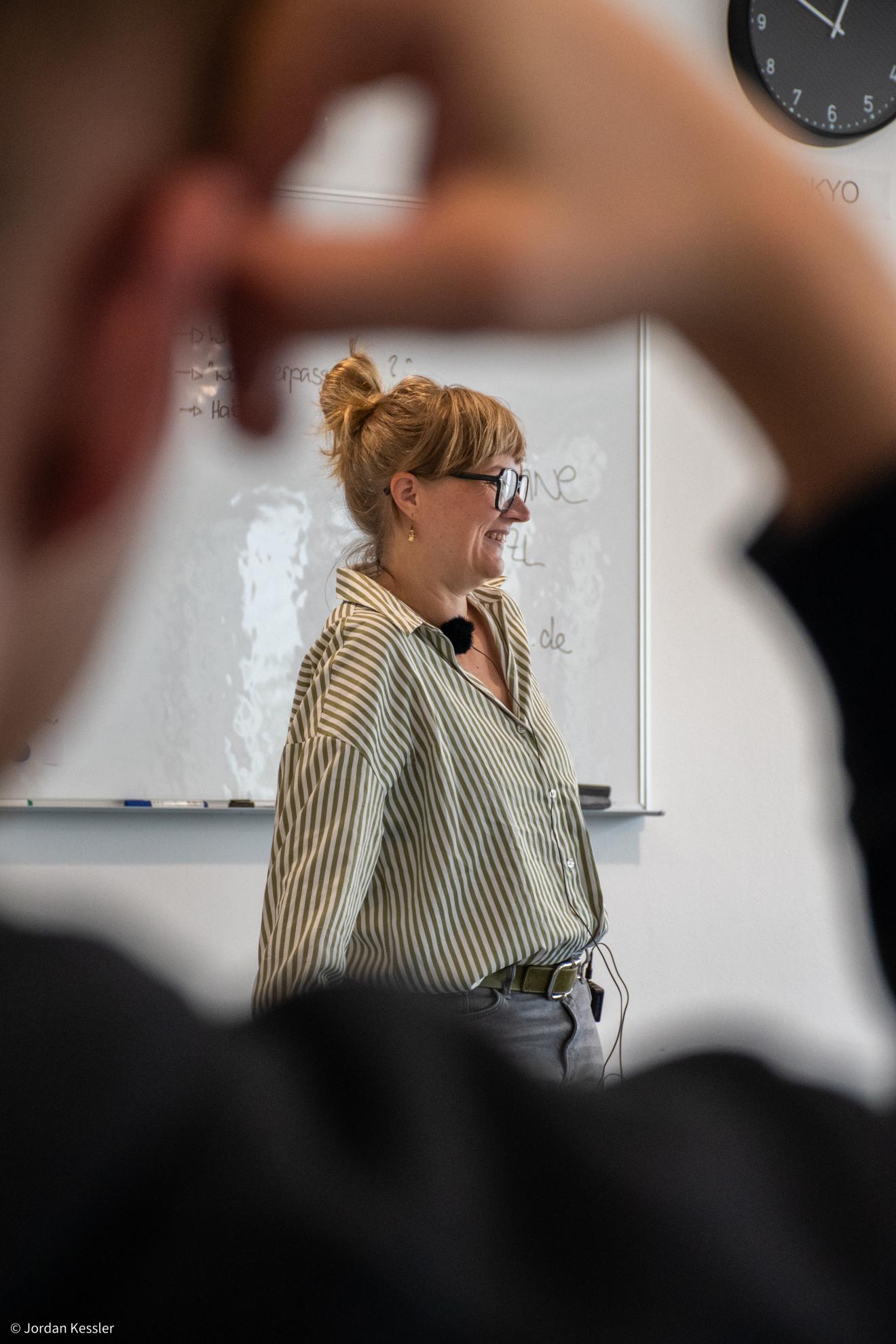 Christine Spatzl steht lächelnd vor einer Whiteboard-Tafel und spricht während eines Workshops. Sie trägt eine gestreifte Bluse und ein Mikrofon, im Vordergrund sind unscharfe Silhouetten von Teilnehmenden zu sehen.