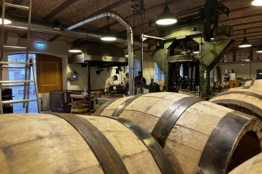 m Vordergrund liegen mehrere fertige Holzfässer, dahinter arbeiten Menschen an Werkbänken zwischen großen, grünen Maschinen. Die Halle verfügt über eine gewölbte Ziegeldecke und industrielles Licht und vermittelt eine handwerkliche Arbeitsatmosphäre.