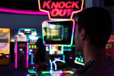 Ein junger Mann steht in einer dunklen, bunt beleuchteten Spielehalle und blickt auf einen grell leuchtenden Spielautomaten mit der Aufschrift „KNOCK OUT“. Die Szene ist farbenfroh, mit vielen Neonlichtern im Hintergrund.