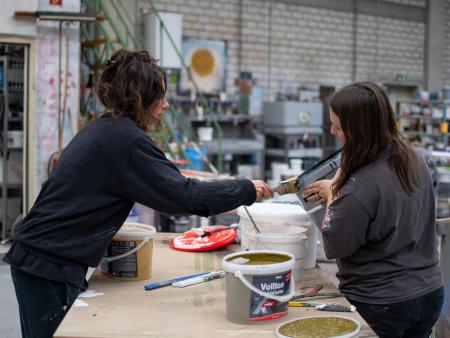Zwei Bühnenmalerinnen bereiten eine neue Farbmischung vor. Eine hält den Eimer, die andere reinigt mit dem Pinsel den Rand und zeigt dabei den sorgfältigen Umgang mit Material und Farbe.