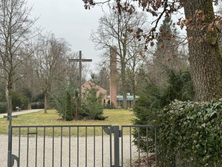 Eingang eines Friedhofs. Es ist leicht links eine großes Kreuz zu sehen und im Hintergrund die Trauerhalle