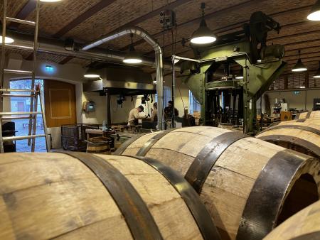 m Vordergrund liegen mehrere fertige Holzfässer, dahinter arbeiten Menschen an Werkbänken zwischen großen, grünen Maschinen. Die Halle verfügt über eine gewölbte Ziegeldecke und industrielles Licht und vermittelt eine handwerkliche Arbeitsatmosphäre.